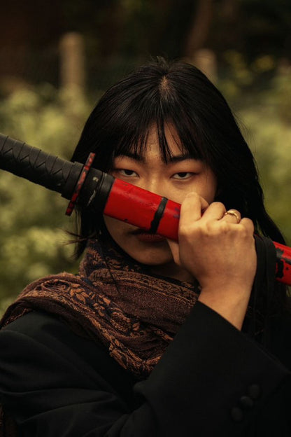 Close-up of an illustrated woman with black hair holding a red and black katana across her face, showcasing intense eyes and a fierce expression. The design reflects bold and artistic streetwear inspiration, perfect for modern fashion enthusiasts.